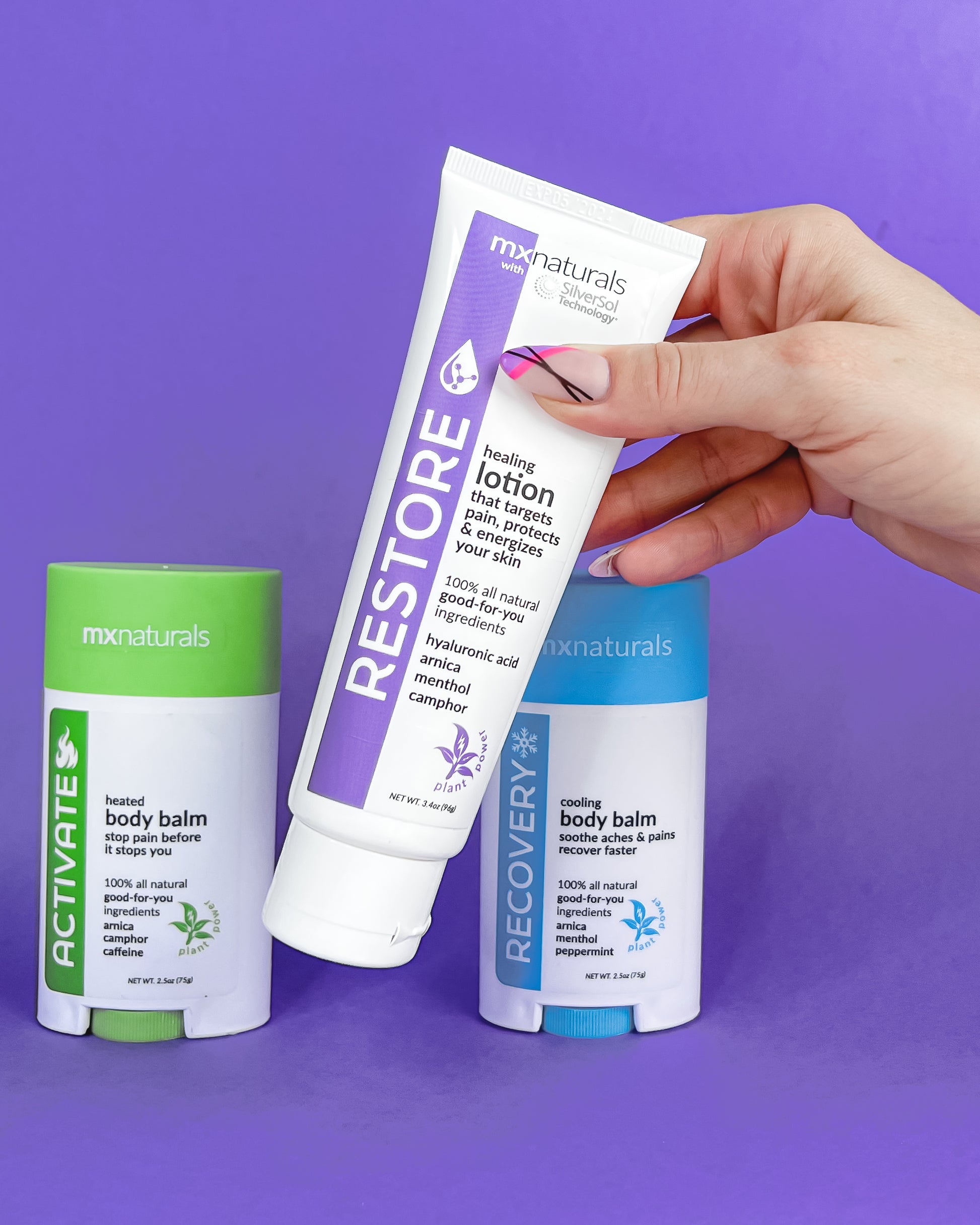 well manicured caucasian female hand picking up a white and purple "restore healing lotion that targets pain, protects and energizes your skin". two additional products, "activate heated pain relief body balm" and "recovery cooling pain relief body balm" rest on the lower plane against a purple background.