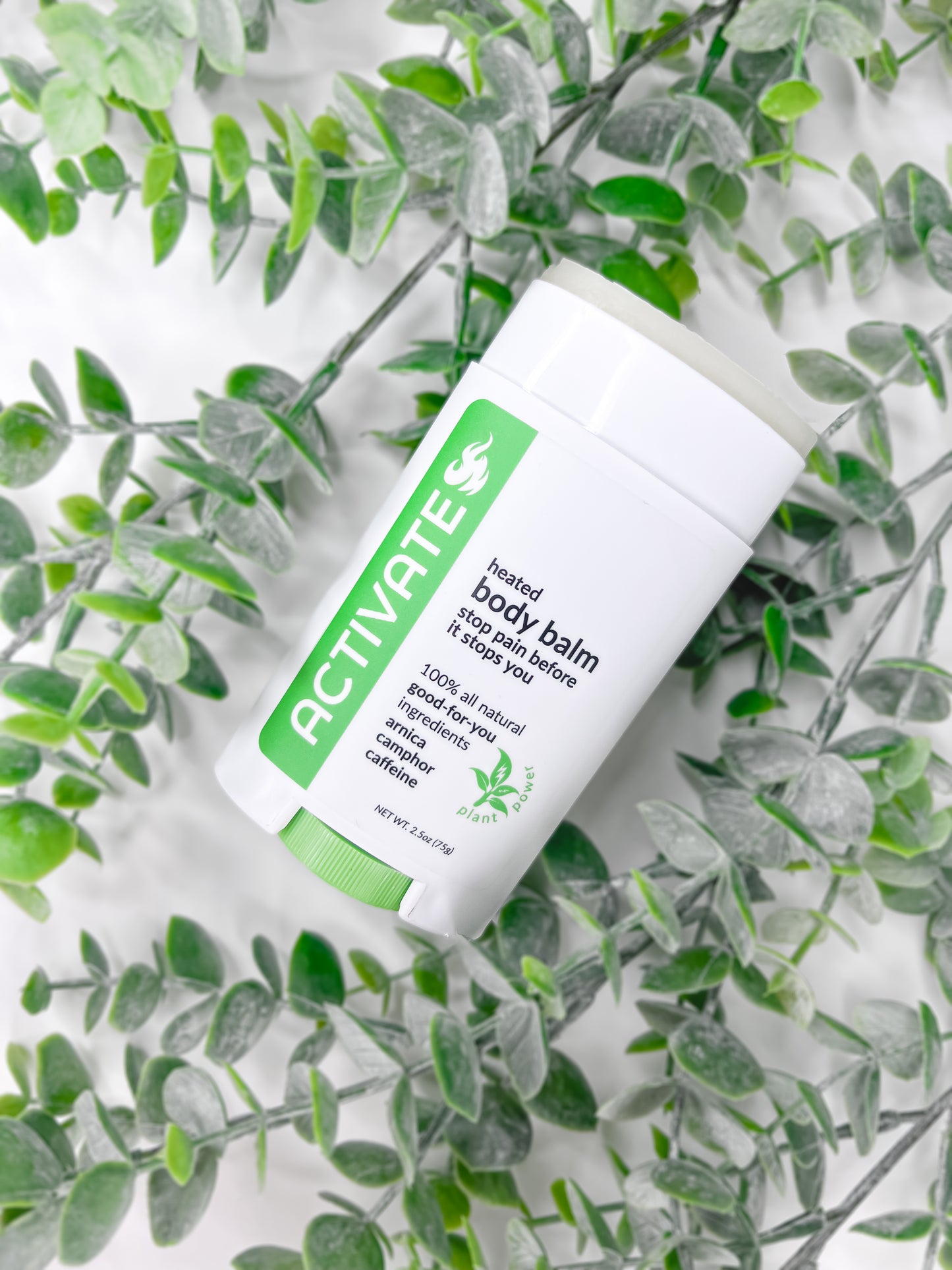 an uncapped white and green activate heated pain relief balm on a white background covered in strands of eucalyptus.