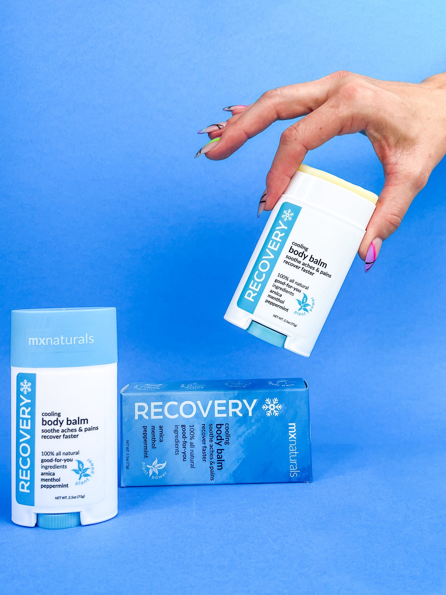 well manicured caucasian female hand picking up an uncapped white and blue "recovery cooling pain relief body care balm"; at a 45 degree angle. two additional products rest on the lower plane against a blue background.