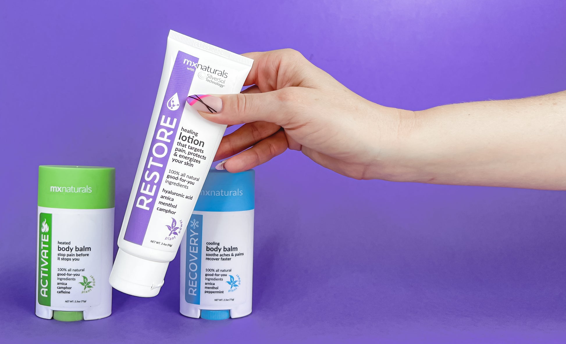 well manicured caucasian female hand picking up a white and purple "restore healing lotion that targets pain, protects and energizes your skin". two additional products, "activate heated pain relief body balm" and "recovery cooling pain relief body balm" rest on the lower plane against a purple background.