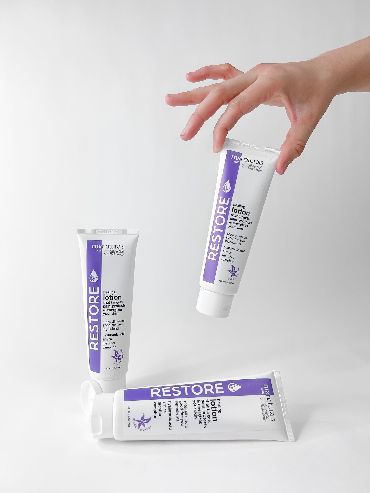 well manicured female hand holding a white and purple lotion tube, "restore targeted pain relief and hydrating lotion" on a white background.